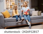 Small photo of Warm Mother's Day moment at home: On a plush sofa, a daughter watches with bated breath as her excited mother delicately removes the bow from her present, their bond evident in their smiles