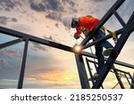 A welder is welding steel on a steel roof truss. Working at height equipment. Fall arrestor device for worker with hooks for safety body harness. Worker in construction site.
