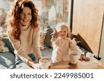 Smiling happy European woman with red wavy hair sitting in cafe in sunny day with her little daughter. Portrait of gorgeous stylish young mom with brown hair and little girl looking away.