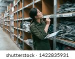 Small photo of asian female warehouse worker looking up the tall shelves doing inventory control. young lady employee in army green shirt uniform count product in stockroom. storehouse staff with clipboard and pen