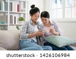 Small photo of Two joyful young excited asian women friends having fun point on mobile phone screen while sitting on comfortable sofa at home. Female friendship chatter funny pictures on cellphone pleasant memory