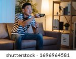 Small photo of cheerful young asian chinese man watching movies on tv at home late in evening. handsome guy relax sitting in comfort couch stay up late at night enjoy television program. male holding remote switch.