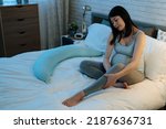 Small photo of smiling asian pregnant woman sitting on bed is massaging her legs to relieve edema before going to sleep in the bedroom at home during nighttime.