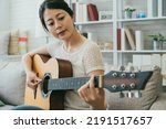 pretty asian Japanese lady in casual wear is plucking the string while learning to play a an acoustic guitar on the couch in a cozy bright living room at home.