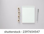 Flat lay of an open blank white page of a notepad with a pen on a side and numbers 2024 on wooden cubes, light lilac background with copy space
