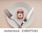 Red alarm clock on white plate with cutlery. Concept of intermittent fasting, lunchtime, diet and weight loss.