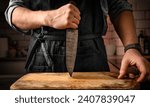Small photo of Chief man in apron holding knife and stubs blade to wooden board at kitchen. Restaurant proffesional with culinary tool