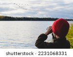 Kid on lake shore watching in binoculars how flock of migrating birds training to fly in wedge