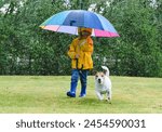 Kid in raincoat under umbrella and her dog playing on grass lawn during spring rain