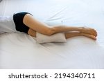 A woman with an anatomical pillow between her legs and knees, lying on a bed with white sheets. A girl resting on her side