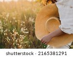 Small photo of Stylish boho woman walking with straw hat in hand close up among wildflowers in sunset light. Atmospheric moment. Summer travel. Young female in rustic linen cloth relaxing in summer meadow