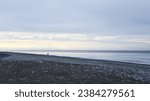 Small photo of A solitary individual is captured in the distance, traversing the desolate, rocky beach that meets the cold blue embrace of the sea, creating a poignant scene of isolation and the vastness of nature