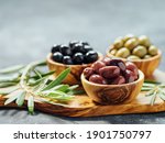 Small photo of Set of green, red and black olives on gray background. Different types of olives in olive wooden bowls and olive oil over wooden cutting board and fresh olive leaves. Copy space.