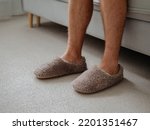 Small photo of Cropped shot of unrecognizable man in soft plush warm slippers, sitting on sofa in morning time at cozy scandinavian home. Hairy legs of adult man in trendy warm slippers on floor