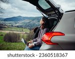 A beautiful young girl is working on a laptop in the trunk of a car and admiring the views of the mountains while traveling through the highlands. Remote work, downshifting, work and travel.