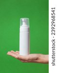 Small photo of Female hand holding cosmetic blank plastic bottle tube jar jolt of face cream, foam. White container on hand on green background. Template mock up for cosmetic product design. Skin care beauty concept