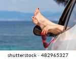 Female legs and hand holding red sunglasses through car window against blue sea background. Travel and summer vacation concepts.