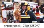 Small photo of Portrait of caucasian handsome fireman in helmet and gull equipment holds girl with teddy bear in arms and looking into camera smiling. The concept of saving lives, heroic profession, fire safety