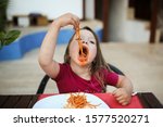 Small photo of Funny grimy girl child eating pasta with tomato sauce in a cafe on the street, poor behavior at the table, ill-bred dirty child. Children's crisis and protest