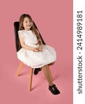 Small photo of A little girl sitting on a black chair on a pink background is very happy and very positive because she is enjoying life. She has a very special dress and very beautiful and arranged hair.