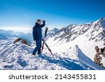 Small photo of St. Anton am Arlberg. March 10, 2022. Young man standing beside skis on top of snowy mountain, Skier stands on the background of beautiful view of snowcapped mountains