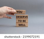 Small photo of You deserve to be well symbol. Wooden blocks with words You deserve to be well. Beautiful grey background. Businessman hand. You deserve to be well concept. Copy space.