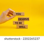 Small photo of You deserve to be well symbol. Wooden blocks with words You deserve to be well. Beautiful yellow background. Businessman hand. You deserve to be well concept. Copy space.