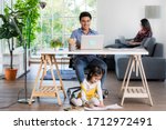 Mixed race family sharing time in living room. Caucasian father using notebook computer to work and half-Thai playing and painting under desk while Asian mother with laptop working her job on sofa.