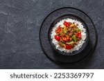 crispy ground beef with stir-fried veggies and a sticky-sweet orange sauce topped on rice in black bowl on concrete table, horizontal view from above, flat lay, copy space