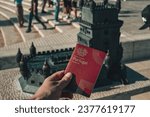 Small photo of Lisbon, Portugal - 03.10.2023: Holding a Portuguese Passport in front of the Belem Tower aka Torre de Sao Vicente miniature model on a sunny summer day.