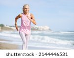 Small photo of Older female doing sport to keep fit. Mature woman running along the shore of the beach. Concept of healthy living in the elderly. Senior woman in fitness clothing running along beach