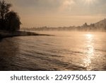 Steam rising from Rhine river near Unkel, Germany in winter as snow and ice covers the landscape with a castle peeking out of the mist and a bird on the river'r edge at golden hour during sunset