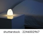 Cute cat shaped night lamp standing on a bedside table next to bed. Bedside lamp. Night lamp standing next to bed. Bedroom lamp on a night table next to a sleeping bed in a dark room. 