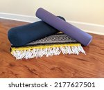 Small photo of Restorative yoga props on a wooden laminate floor with a neutral wall background. Props include a purple yoga mat, a navy bolster, and a patterned blanket.