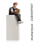 Small photo of Schoolboy sitting on a tall white column and reading a book isolated on white background