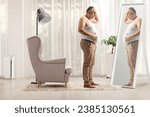 Unhappy mature man holding his big belly and looking at a mirror at home