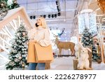 Low-angle view of pretty blonde young woman talking smartphone and holding paper bags with purchase standing in hall of celebrate shopping mall in Christmas eve, on background of xmas decorations.
