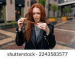 Small photo of Portrait of displeased pretty young woman touching wet hair after autumn rain standing on beautiful city street. Front view of stressed lady untangling hair after being caught in rain.