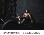 Young dedicated active strong fit sweaty muscular man wit big muscles sitting on the floor of the gym after barbell weight lifting workout and taking a break from hardcore cross training real people