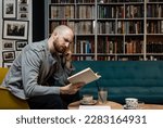Clever serious caucasian male in casual wear reading new interesting novel in bookstore, successful joyful man student holding positive book while sitting near shelves in university library