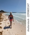 Small photo of Elderly woman with hat and bathing suit walking on tropical beach with sargasso