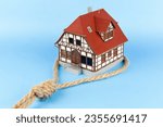 Small photo of Plastic model of a house in a hangman's noose on a blue background. The house is trapped. It is difficult to pay for buying a house on credit.