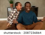 African American laughing dad trying to meditate and keep calm while his misbehaving dark-skinned 9-years-old son screaming in his ear with frustration, demanding absolute attention