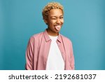 Small photo of Portrait of funny black girl with blond curly hair showing tongue winking with one eye, making faces, fooling around, aping with silly face, joking, teasing, isolated on blue studio background