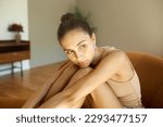 Small photo of Gorgeous sad female in home clothes sitting on couch hugging legs resting chin on knees, looking aside with pensive facial expression, missing her ex-boyfriend. Human emotions and feelings
