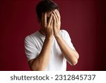 Small photo of Portrait of man looking thorough fingers covering face with palms, feeling shy and embarrassed standing against red studio background. People and emotions, signs, gestures, body language concept