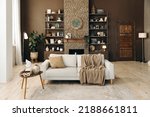 Modern stylish living room with large windows and beige sofa on the background of brown wall with fireplace, shelving with books and decor, and potted plants. Cozy chalet interior. Empty space.