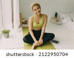 Small photo of Toothy smiling African teenage blonde with curly shortcut in sportswear sitting on yoga mat in white stylish minimalistic room, legs crossed, pad with blank copy space for your advertisement on floor