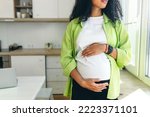 No face picture of pregnant african American woman with long curly hair looking through window standing in kitchen, rubbing her big cute belly, expecting baby boy to be born in few month