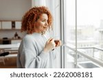Side view indoor portrait of young attractive woman with red curly hair looking through window holding transparent cup of hot tea, admiring winter cityscape, dressed in stylish sweater